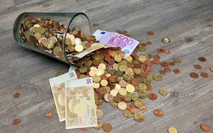A glass jar with euro coins and banknotes spilled on a wooden surface, representing finance and savings.