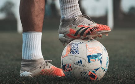 Close-up of a soccer player wearing Adidas cleats, holding a branded soccer ball on grass.