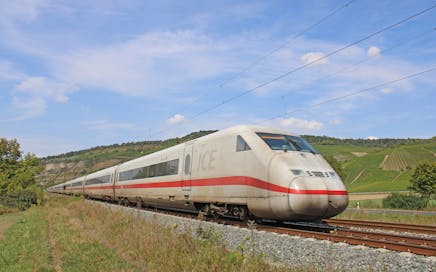 A high-speed train travels through a scenic rural countryside with green fields under blue skies.