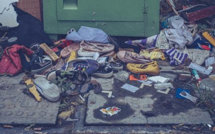A recycling bin surrounded by clothes and litter on a city street, highlighting waste management issues.