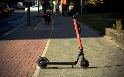 A Voi e-scooter parked on a sunlit city sidewalk with a blurred urban background.