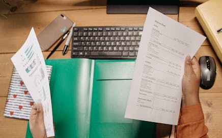 Hands holding and reviewing balance sheets over a desk with a keyboard and stationery.